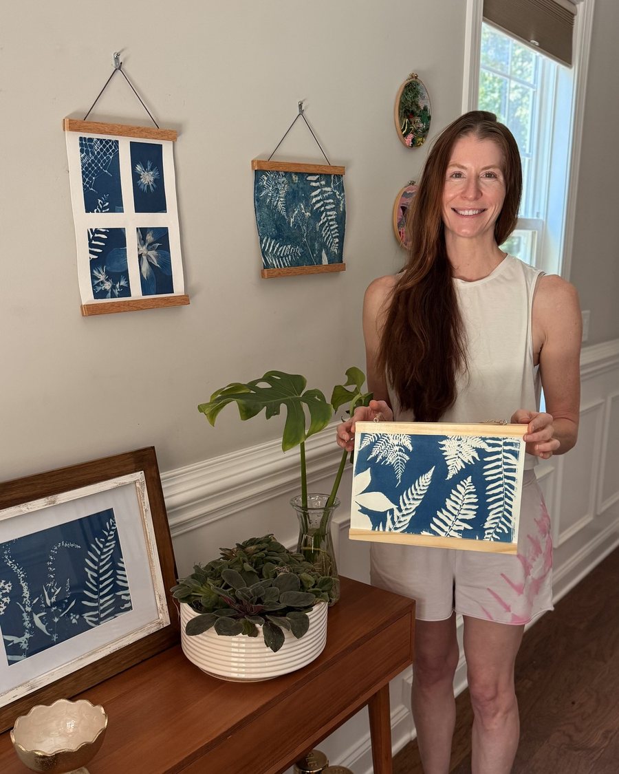 Kristi smiling and holding a framed ferns cyanotype print, standing beside a console table with more of her framed prints on the wall behind her