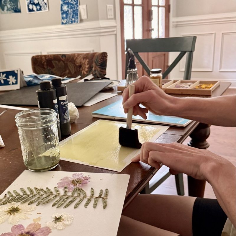 Module 2: Kristi brushing pale yellow-green sensitizer onto watercolor paper with a foam brush, pressed ferns and flowers waiting beside it
